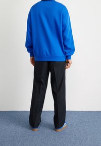 Sininen collegepaita, jossa rento istuvuus, resorit hihoissa ja helmassa. Mustat suoralahkeiset housut. Yksinkertaiset siniraidalliset sandaalit teksturoidulla lattialla.