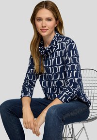 Frau mit langen, hellbraunen Haaren, die ein marineblau gemustertes Oberteil mit Rollkragen und dunkelblaue Jeans trägt, sitzt auf einem Metall-Drahtstuhl.