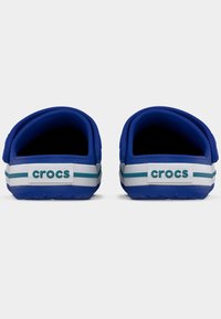 Ζευγάρι μπλε και λευκά παπούτσια Crocs από την πίσω όψη με τυρκουάζ ρίγες και λαστιχένιους ιμάντες με λογότυπο στη φτέρνα, πάνω σε απλό φόντο.