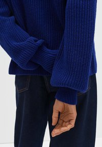 Pull tricoté bleu profond avec un motif côtelé texturé. Manches larges et poignets au niveau des poignets, associé à un pantalon en denim foncé.