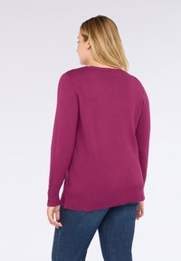 Mujer con cabello rubio lleva un suéter de manga larga magenta y jeans azules, de espaldas frente a un fondo gris claro.