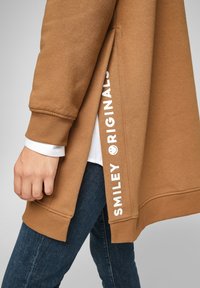 Sudadera marrón con puños acanalados, abertura lateral y logo "SMILEY ORIGINALS" en blanco. Lleva una camisa blanca debajo y está combinada con pantalones vaqueros oscuros.