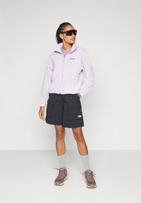 Heled lilla fliisjakke, mustad cargo-shorts, millel on plaastrid, hallid lühikesed sokid ja pruunid matkasaapad. Mudel, kes kannab prille, seisab poosis.