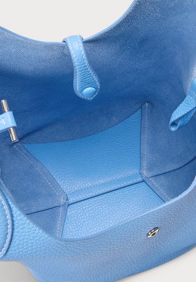 Interiér modré kožené tašky, který má texturovaný povrch, hladký lem a malý stříbrný kovový detail, s rozdělenými přihrádkami pro organizaci.