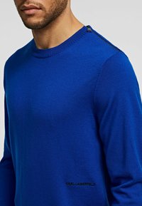 Blauer gestrickter Pullover mit rundem Hals, gerippten Bündchen und Schulterdetails aus Metall. Das Logo "KARL LAGERFELD" ist am Saum gestickt.
