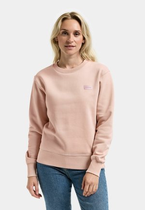 Jasnoróżowa bluza z okrągłym dekoltem i ściągaczami na mankietach. Posiada małą naszywkę z logo na górze po lewej stronie. W zestawie z dżinsami.