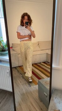 Bílý tričko a žluté květinové pyžamové kalhoty s volným střihem, stojící na dřevěné podlaze. Přítomné zrcadlo, neformální vnitřní prostředí.