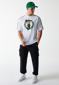 Camisa blanca de malla de manga corta con el logo de los Boston Celtics, pantalones cargo negros y zapatillas blancas, complementados con una gorra verde.