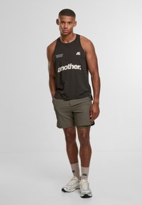 Canotta nera senza maniche con testo grafico bianco, abbinata a pantaloni corti verde oliva. Presenta scarpe da ginnastica chiare e calzini beige.