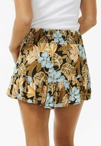 Frau trägt einen schulterfreien weißen Oberteil zu einem kurzen Rock mit floralem Muster, der braune, blaue und beige Blätter und Blumen zeigt.