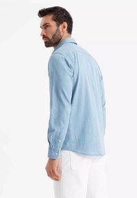Chemise bleu clair à manches longues en tissu doux, avec un ourlet arrondi. Le modèle porte un pantalon blanc.