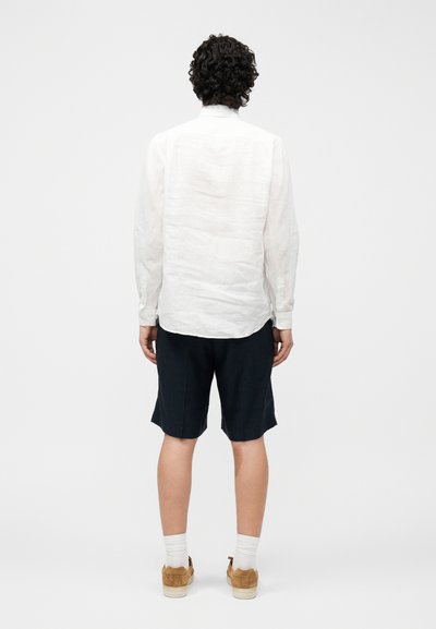 Άτομο με σγουρά σκούρα μαλλιά στέκεται με την πλάτη, φοράει λευκό πουκάμισο με μακριά μανίκια, μαύρο σορτς, λευκές κάλτσες και καφέ παπούτσια σε απλό φόντο.