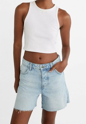Femme porte un top court blanc sans manches et un short en jean bleu clair aux bords effilochés, debout, une main dans la poche.