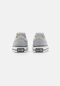 Une paire de baskets Converse All Star basses gris clair montrées de derrière sur un fond blanc avec des détails de semelle en noir et blanc.