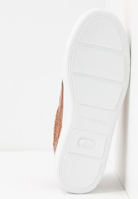 Semelle en caoutchouc blanc avec grip texturé et logo en relief, fixée à une chaussure en cuir marron à motif crocodile.