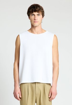 Uomo con capelli castani corti che indossa una canottiera bianca e pantaloni beige su uno sfondo chiaro e semplice.