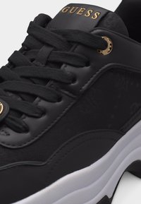 Baskets noires Guess avec lacets noirs, œillet doré marqué et logo doré "GUESS" sur la languette, avec une semelle blanche.