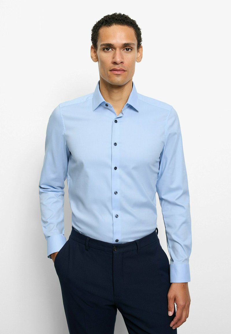 Light blue dress shirt with long sleeves, buttoned collar, and black buttons. Worn with dark pants, featuring a smooth texture and tailored fit.