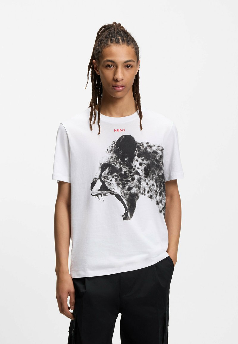 T-shirt en coton blanc avec un graphisme léopard noir et le texte "HUGO" rouge au-dessus. Manches courtes et coupe décontractée, complété par un pantalon foncé.