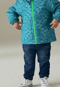 Jacket turchese a patttern con disegni grafici, zip verde, abbinato a jeans denim blu scuro e sneakers bianche.