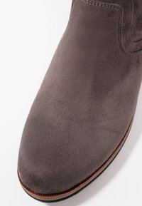 Gros plan sur l'avant d'une botte en daim gris avec des coutures visibles et un bord brun sur la semelle, sur fond blanc.