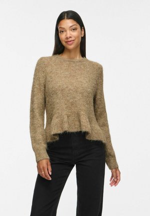 Femme aux longs cheveux foncés portant un pull pelucheux marron clair avec un ourlet à volants et un pantalon noir taille haute, debout devant un fond blanc.