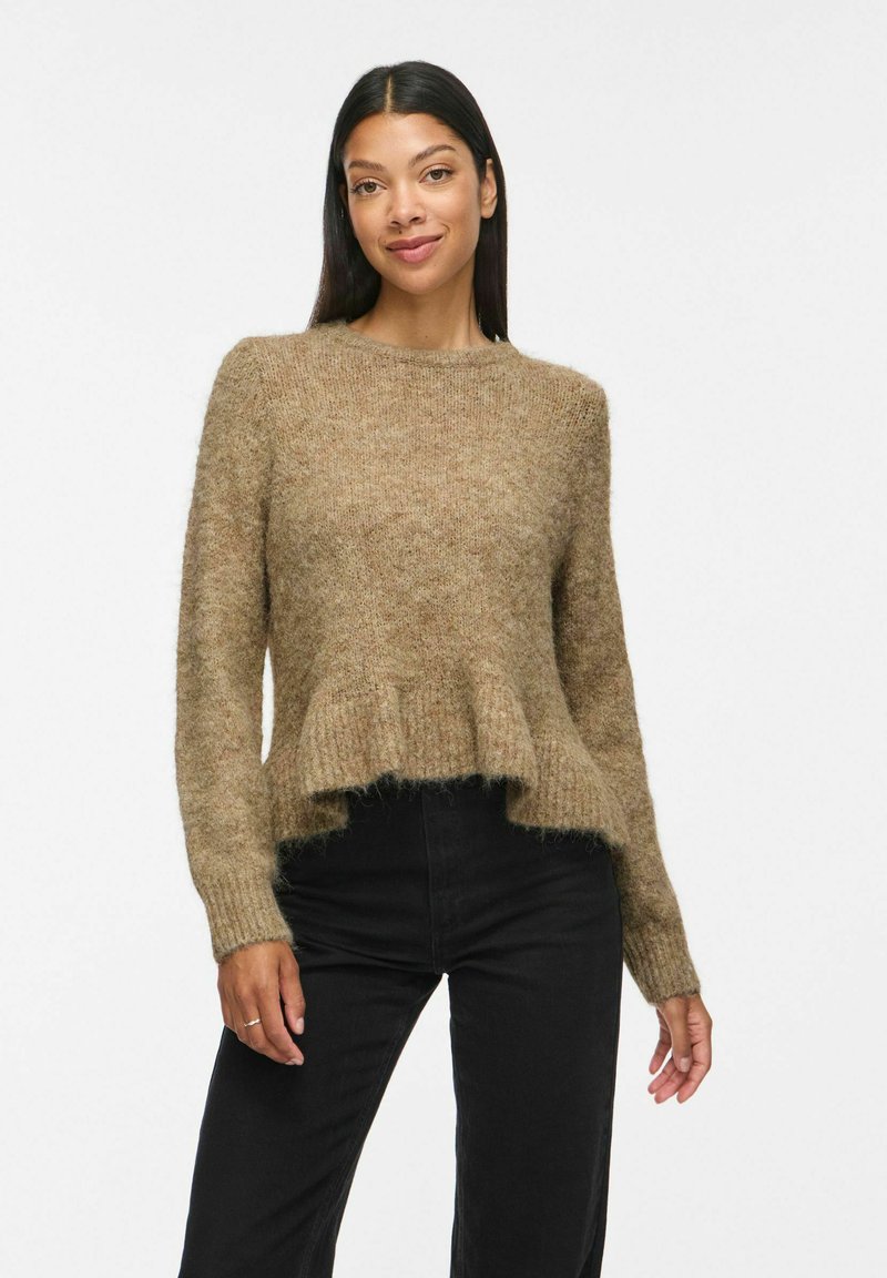 Femme aux longs cheveux foncés portant un pull pelucheux marron clair avec un ourlet à volants et un pantalon noir taille haute, debout devant un fond blanc.