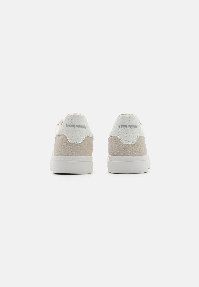 Baskets en daim beige avec des accents blancs, à forme arrondie, col de cheville rembourré et détails perforés. Logo "le coq sportif" affiché à l'arrière.