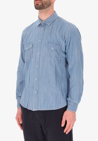 Camicia di denim blu chiaro con colletto a bottoni, due tasche sul petto e maniche lunghe con polsini abbottonati. Vestibilità pulita e dritta.