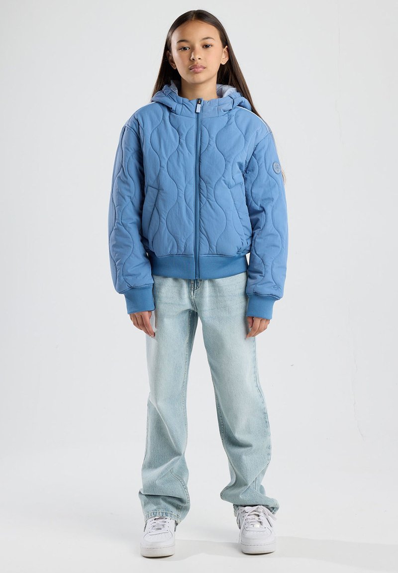 Blue quilted jacket with a hood, featuring a curved pattern, front zipper, and ribbed cuffs. Paired with light blue jeans and white sneakers.