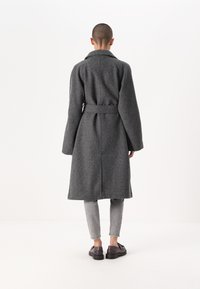 Siv plašč iz volne z pasom, ki ima dolg silhuet, široke rokave in zarezan ovratnik. Nosijo ga čez svetlosive hlače in temne čevlje.