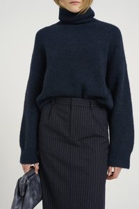 Granatowy sweter z dzianiny z golfem o teksturowanym wykończeniu i szerokimi rękawami, zestawiony z ołówkową spódnicą w prążki. W ręku trzymana jest szara kopertówka.