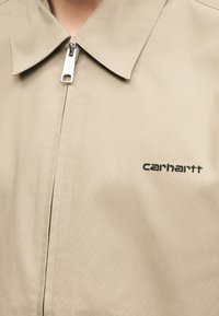 Μπεζ ζακέτα με φερμουάρ, με μυτερό γιακά και μικρό μαύρο κεντητό λογότυπο "Carhartt" στο αριστερό στήθος.
