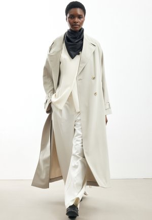 Sort kvinde går, iført en lang beige trenchcoat, hvid dragt, sort tørklæde og sorte sko mod en ensfarvet hvid baggrund.
