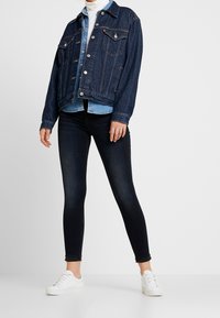 Chaqueta vaquera con un diseño clásico, color azul oscuro, botones plateados y bolsillos en el pecho, combinada con una camisa azul claro y unos vaqueros oscuros.