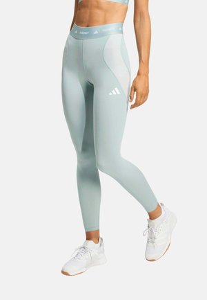 Mujer con mallas Adidas Techfit de color azul claro y zapatillas blancas Adidas, de pie con una pierna ligeramente doblada.