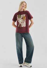 Jonge vrouw die glimlacht, gekleed in een bordeauxrood T-shirt met de tekst "Happy Smile Good" en smiley gezichten, losse blauwe jeans en witte sneakers.