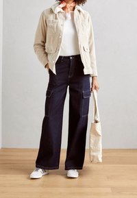 Chaqueta de pana beige con forro de piel, camisa blanca, jeans cargo oscuros y zapatillas blancas. Sosteniendo una bolsa de mano crema. Fondo de suelo de madera.