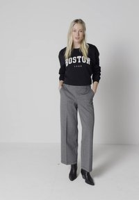 Femme debout les mains dans les poches portant un sweat Boston noir, un pantalon gris à jambes larges et des bottines noires sur un fond uni.