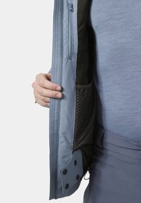 Hånd, der holder en åben lyseblå jakke, der viser en indvendig lodret lynlåslomme og en sort netlomme, båret over en blå tekstureret skjorte og bukser.