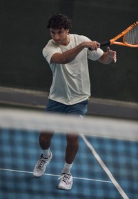 Tennis player in light gray shirt and blue shorts, holding an orange racquet. White sneakers with gray accents. Indoor court setting.