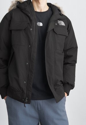 Personne portant une veste isolante noire avec une capuche en fourrure et un t-shirt noir, les deux arborant le logo The North Face. Mains dans les poches.