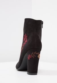 Stivaletto alla caviglia in suede nero con tacco alto, decorato con ricami floreali rosa e beige e una zip sul retro, presentato su uno sfondo bianco.