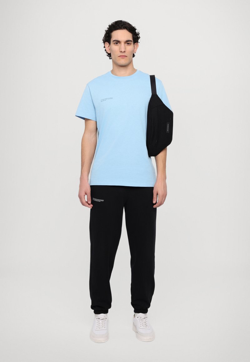 Lyseblå T-shirt, sorte joggers, sort slingtaske, hvide sneakers; enkel design, afslappet pasform, tekstdetaljer på T-shirten, glat stof.
