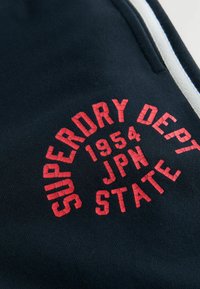 Námořnické tepláky s texturovaným červeným kulatým logem, na němž je výrazně vytištěno "SUPERDRY DEPT 1954 JPN STATE" vpředu.