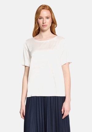 Junge Frau mit rotem Haar, die ein einfaches weißes Kurzarmshirt und einen dunkelblauen plissierten Rock trägt, steht vor einem weißen Hintergrund.