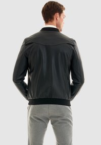 Schwarze Lederbomberjacke mit geripptem Saum und Kragen. Glatte Textur, minimale Nähte und geschwungene Passe auf der Rückseite.