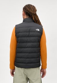 Czarna kamizelka puchowa z wysokim kołnierzem i poziomymi szwami, zestawiona z pomarańczową koszulą z długim rękawem, noszona na jasnozielonych spodniach.