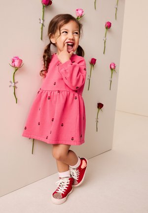 Młoda dziewczyna w różowej sukience i czerwonych trampkach stoi uśmiechnięta obok ściany ozdobionej przyklejonymi różowymi i czerwonymi różami.