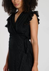 Schwarzes Wickelkleid mit Rüschendetails an den Ärmeln, aus strukturiertem Stoff mit Wellenmuster. Mit einer Bindung in der Taille.